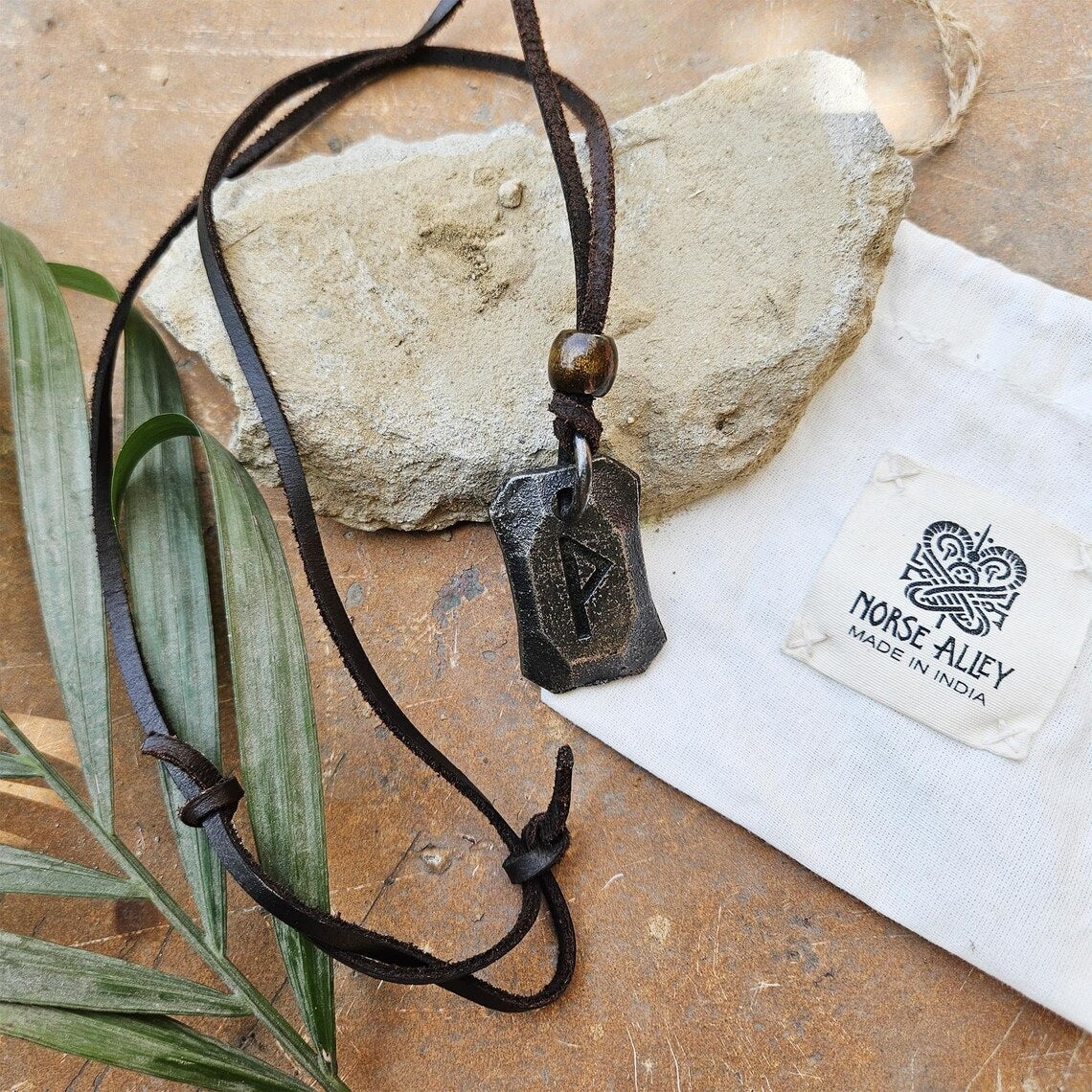 The Viking Rune Pendant Wunjo features a bold geometric rune on a black leather cord, displayed on rock with a green leaf. A branded Norse Alley cloth bag (Made in India) sits beside this striking Norse mythology necklace.