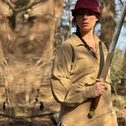 Outdoors in sunlight, a person in a tan Warrior Shirt – Medieval Strength & Style – Unisex and red hat holds a sword with both hands, evoking Norse costume traditions.