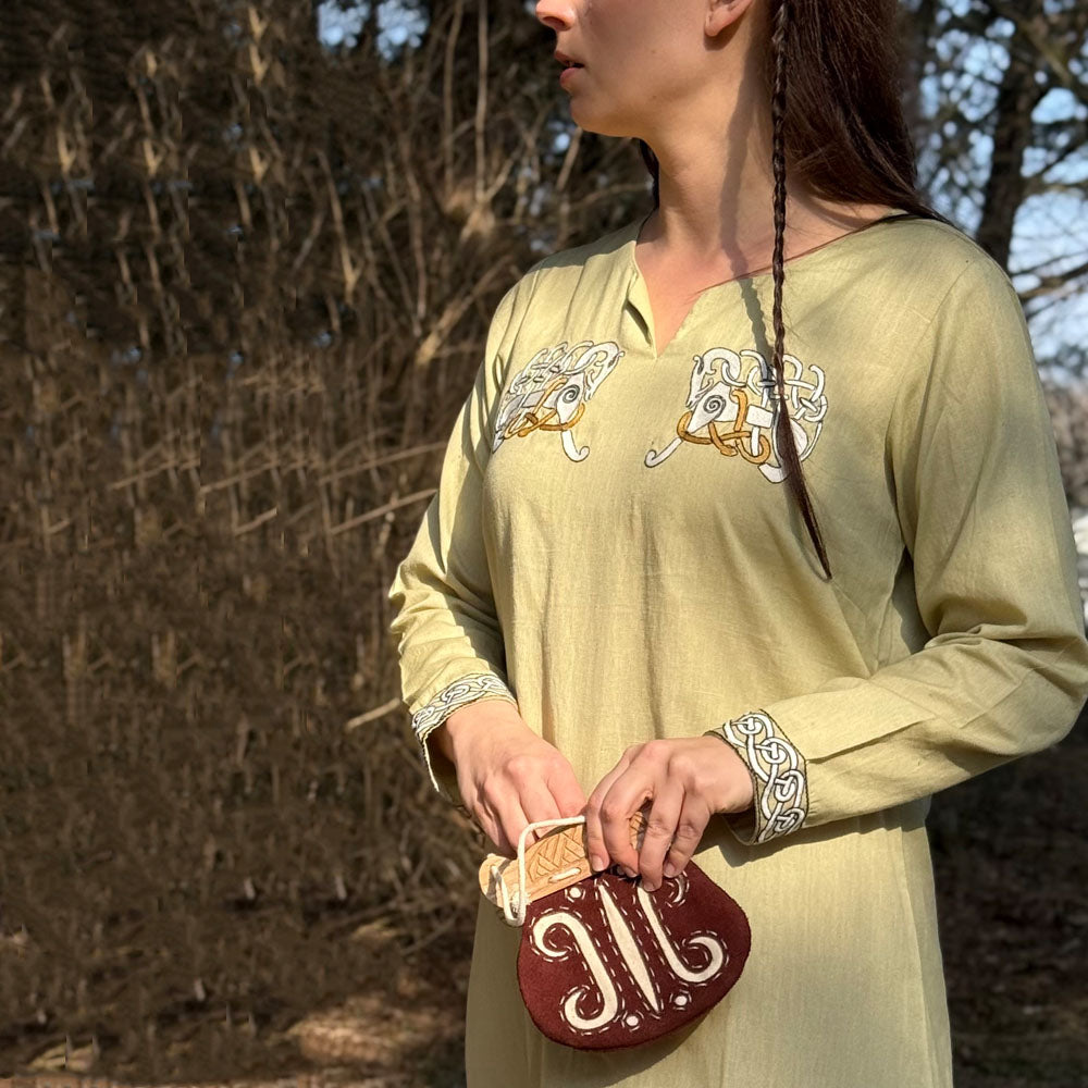 A woman wears the Sleipnir Viking Dress- Ladies in light green cotton-linen with embroidery, standing outdoors with a brown pouch. Her hair is partially braided, and leafless trees are visible in the background.