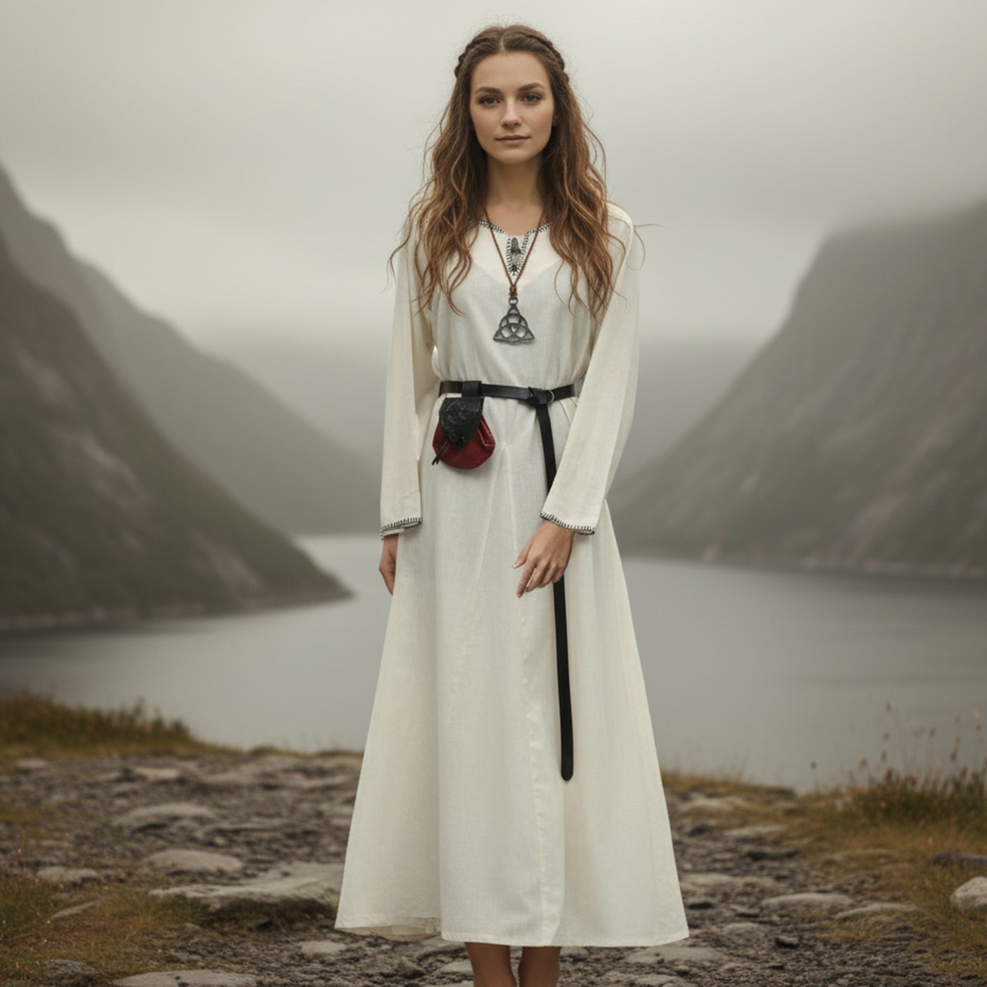 A woman in the Viking Thorndale Dress- Ladies stands on rocky ground by a misty lake, surrounded by steep green mountains under a cloudy sky.