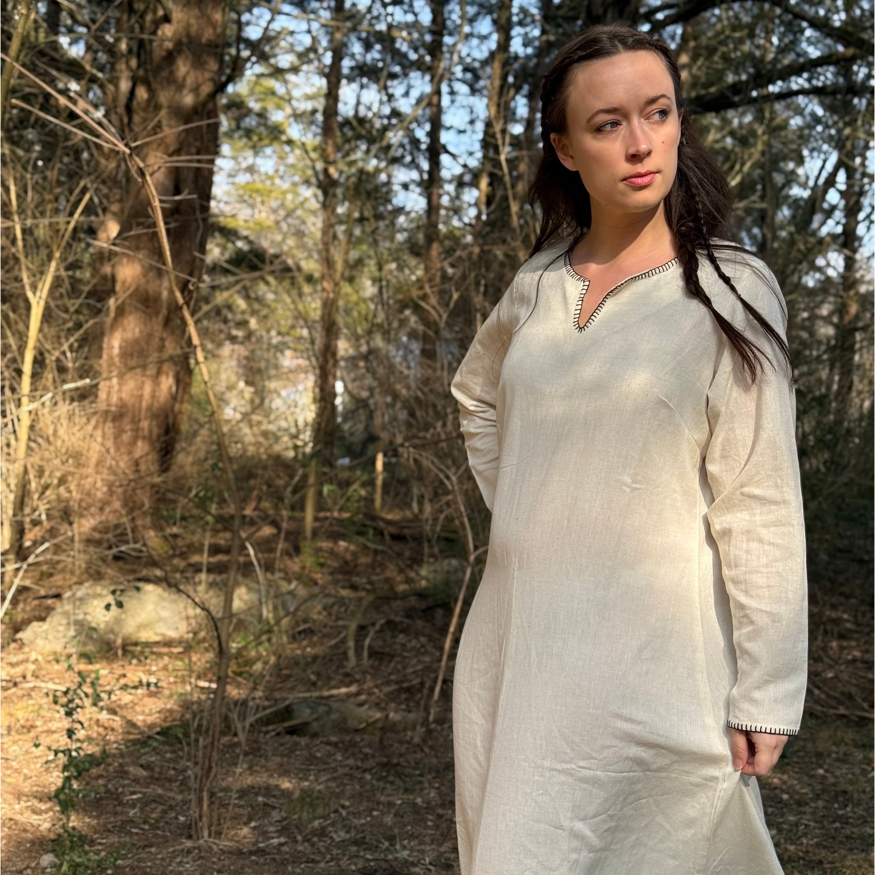 A woman in the Viking Thorndale Dress- Ladies stands in a sunlit forest, gazing to the side with a thoughtful expression as sunlight filters through the trees and casts dappled shadows on the ground.