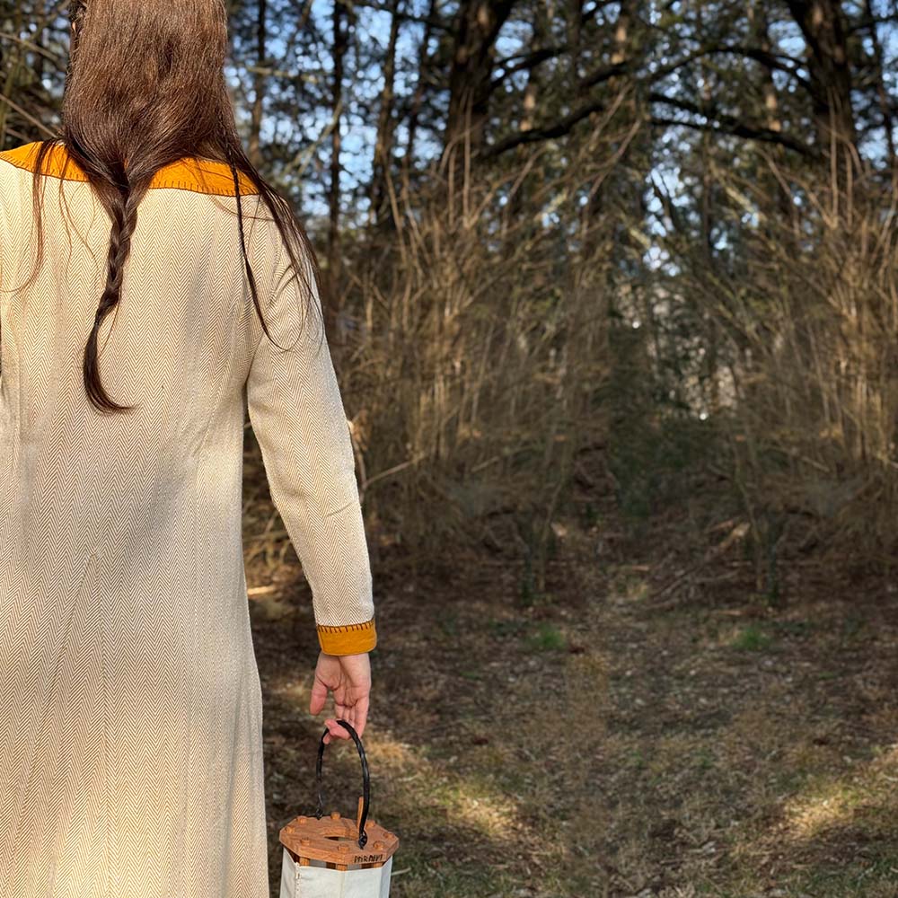 A woman with long brown hair in a braid wears the Nordic Runeweave Viking Dress-Ladies, featuring a cream color and orange trim with Norse-inspired details, standing outdoors holding a lantern amid dry trees and brush.