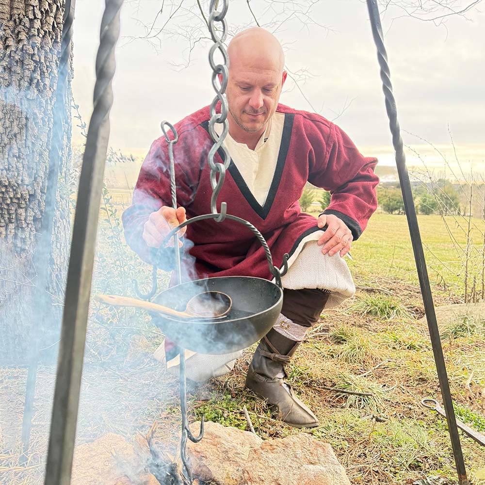 A man in a Viking Woolen Tunic - Men's kneels outdoors, tending a pot over a campfire as smoke rises in a grassy, tree-lined clearing beneath cloudy skies.