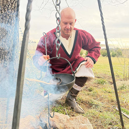 A man in a Viking Woolen Tunic - Men's kneels outdoors, tending a pot over a campfire as smoke rises in a grassy, tree-lined clearing beneath cloudy skies.