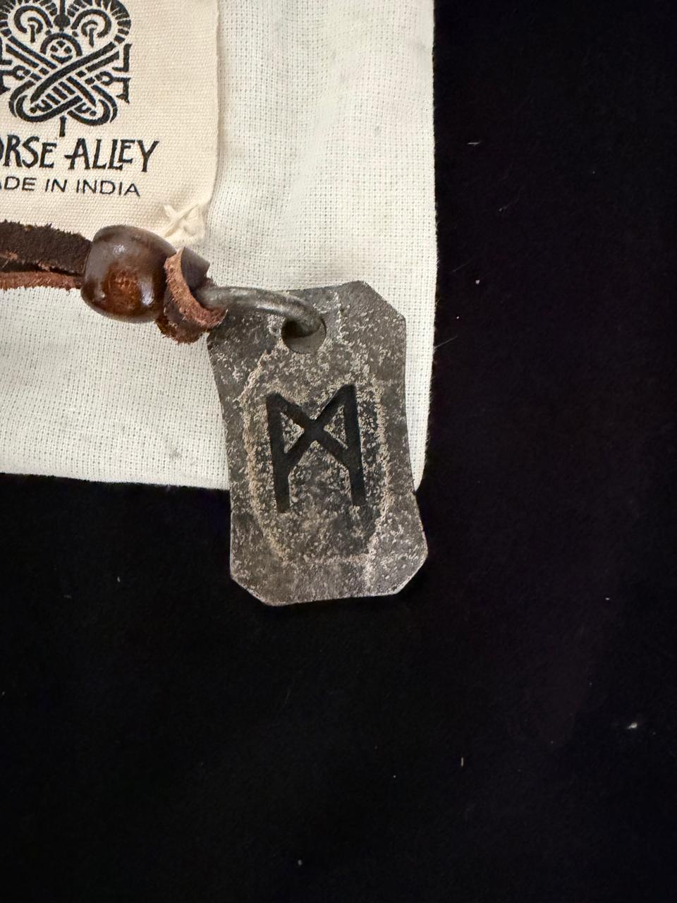 The Viking Rune Pendant Mannaz, crafted from metal, hangs on a leather cord beside a NORSE ALLEY MADE IN INDIA cloth label, all set against a black background.