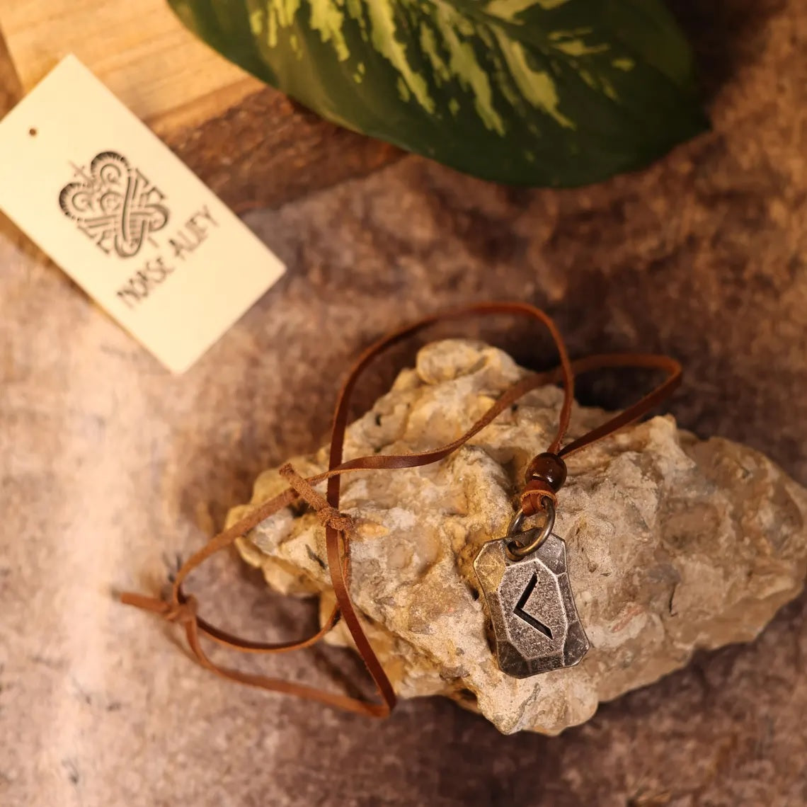 The Viking Rune Pendant Kenaz, a brown leather Norse necklace with a silver Kenaz rune, rests on rough stone beside a Norse Alley tag and green leaf, evoking Norse mythology on a rustic brown surface.