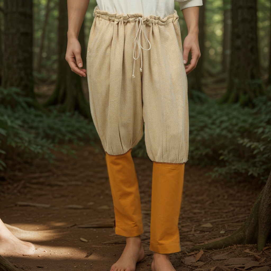 A barefoot man stands on a forest floor in beige Nordic Nomad Viking Pants over slim orange pants, the woodland backdrop evoking the spirit of a Nordic reenactment costume.