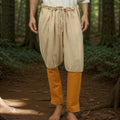 A barefoot man stands on a forest floor in beige Nordic Nomad Viking Pants over slim orange pants, the woodland backdrop evoking the spirit of a Nordic reenactment costume.