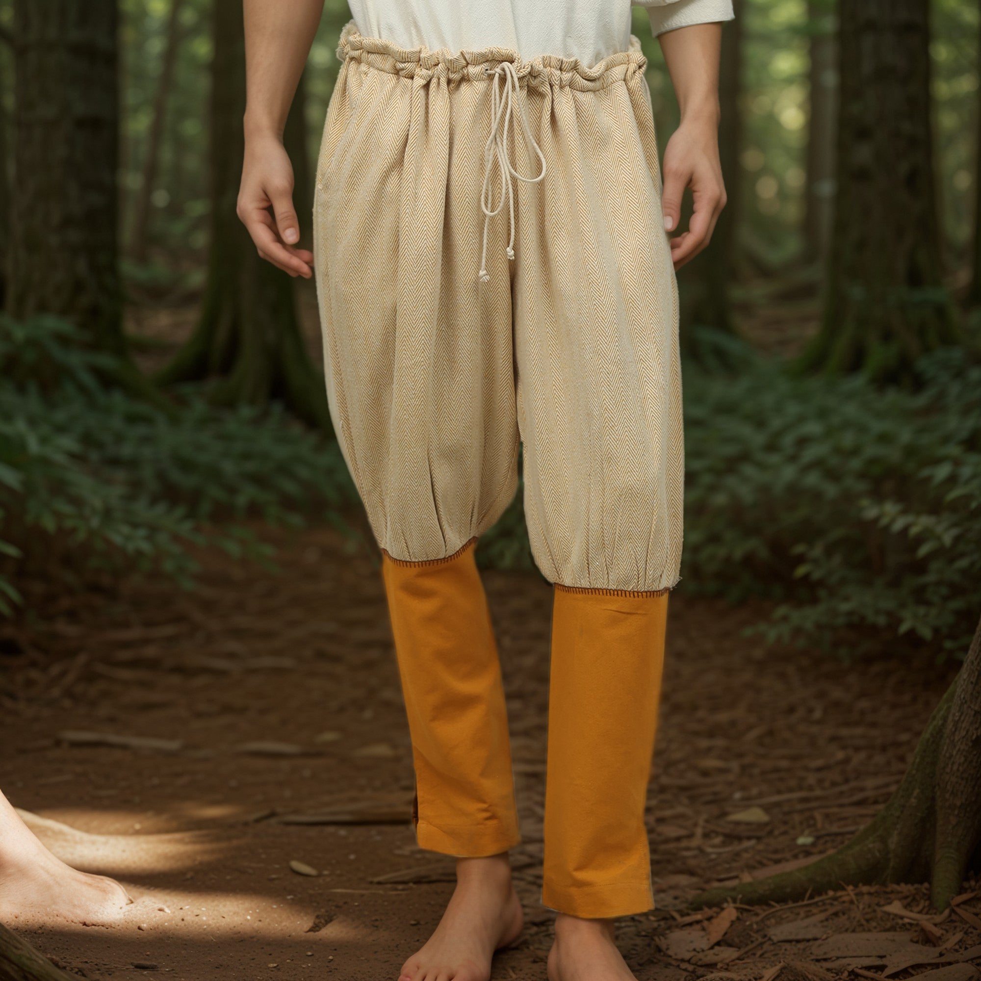 A barefoot person stands in a forest, wearing loose, beige drawstring shorts over Nordic Nomad Viking Pants for men. The scene is lush with trees and greenery, while their upper body is partially out of frame.