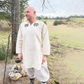 A man stands outdoors by a fire pit wearing Odin's Raven Tunic for men—a cream-colored tunic with black designs near the neckline—surrounded by grass, trees, and a cloudy sky.