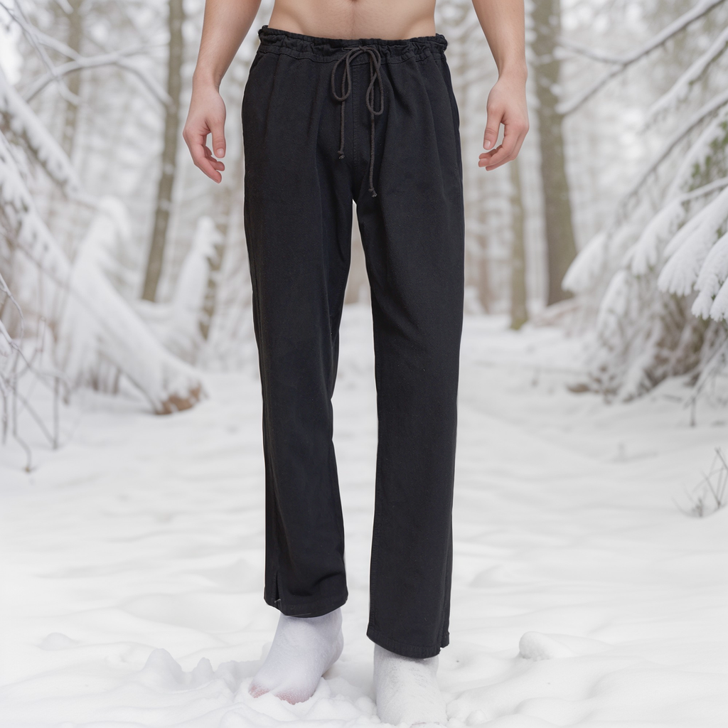 A person stands barefoot in the snow wearing Warrior Pants - Men's; their upper body is out of frame. Snow-covered trees and branches surround them, creating a wintry reenactment forest scene.