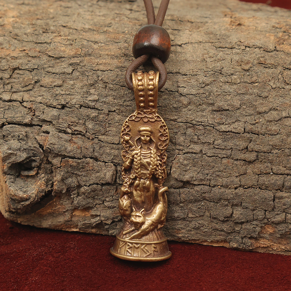 The Freyja Pendant – Goddess of Love and War is a bronze jewelry piece with Norse-style engravings, featuring a bearded figure amid animals on a leather cord with a wooden bead, displayed on rustic bark against vibrant red fabric.