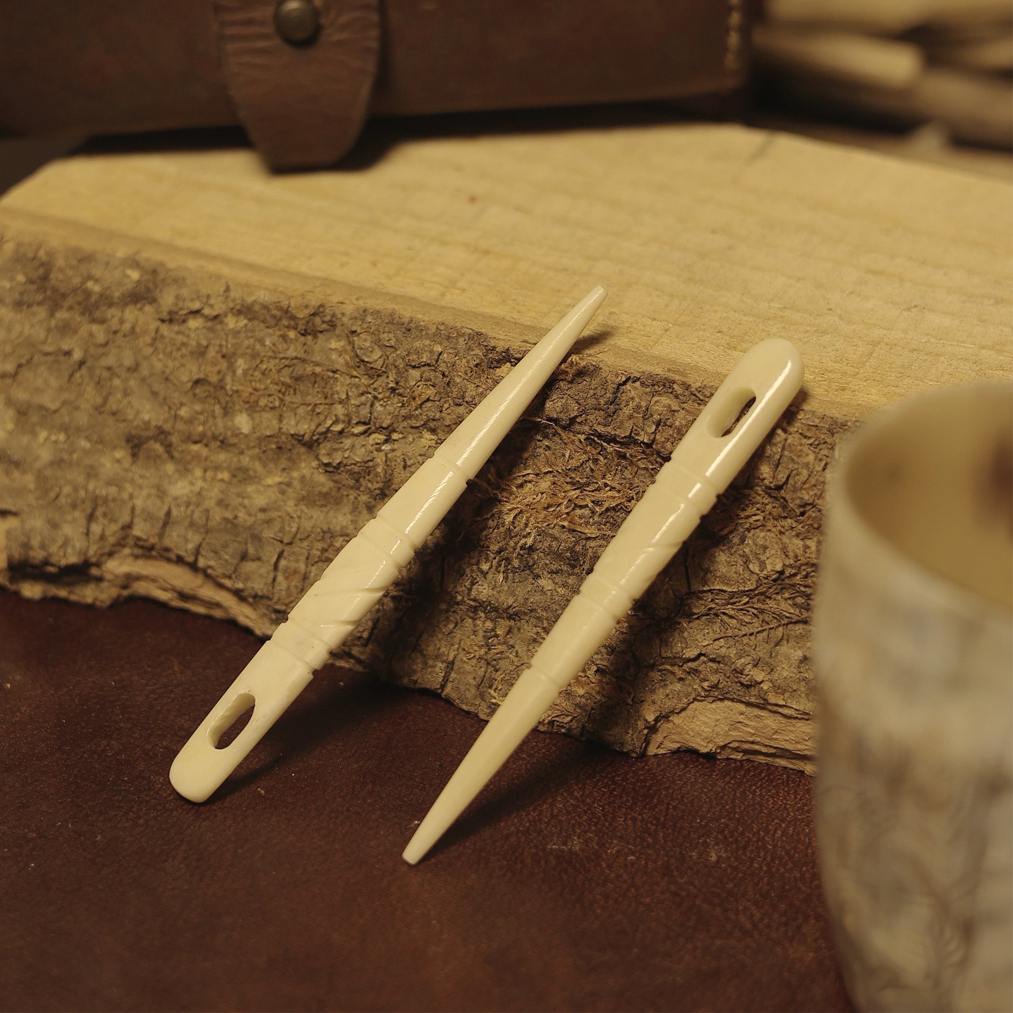 A pair of ivory Bone Knitting Needles – handcrafted in Viking style – rests on rough wood, with a cup and leather case in the background, evoking a rustic, vintage feel reminiscent of traditional knitting tools.