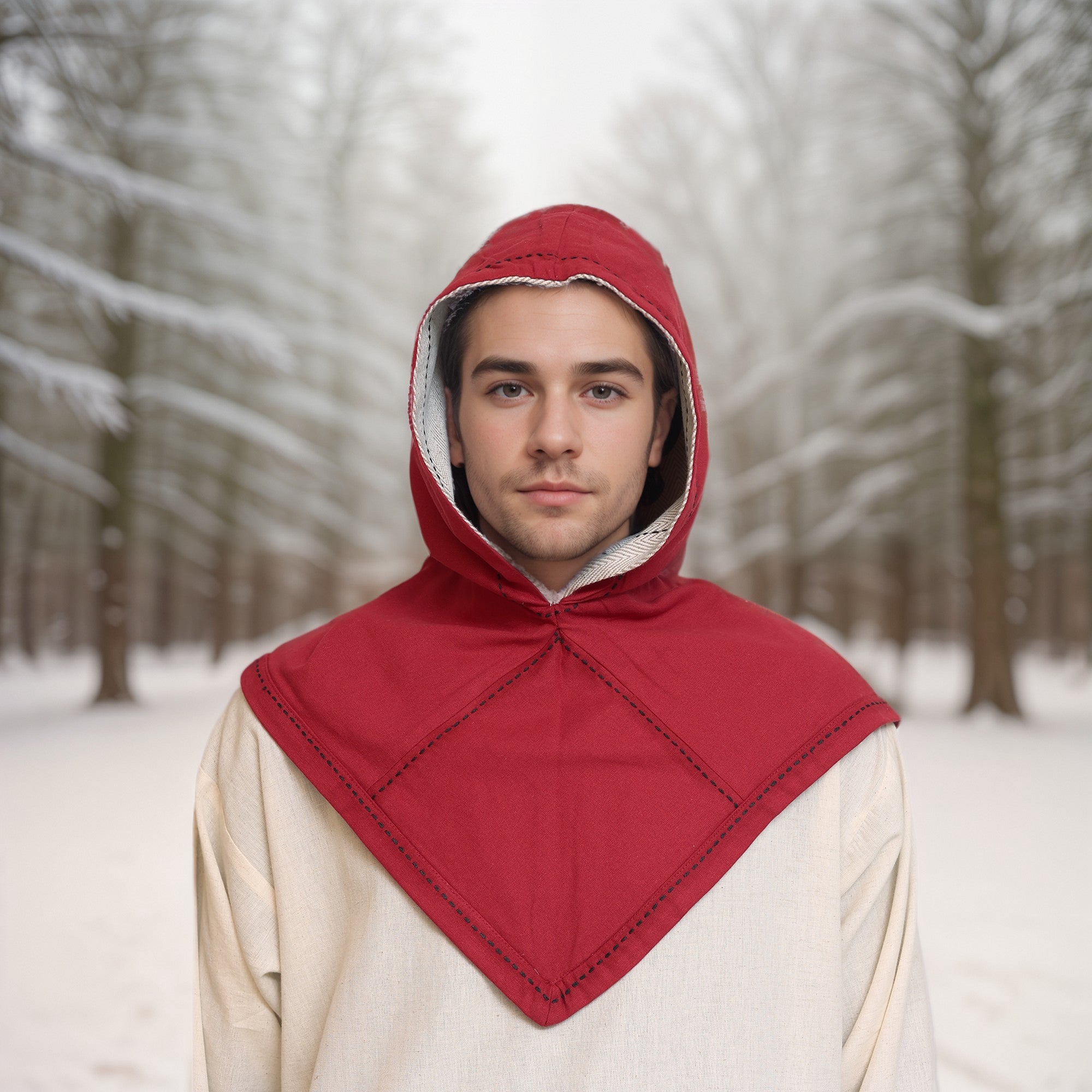 A person in a Reversible Viking Hood, Skjoldehamn, stands in a snowy forest with bare trees. They look directly at the camera, creating a calm, wintry scene.
