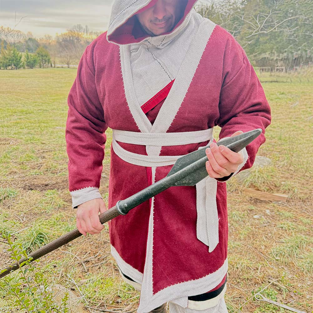 Outdoors, a man wears the Viking Wool Coat-Men's in red and gray with a hood, holds a spear, and stands on grassy ground with trees behind him.