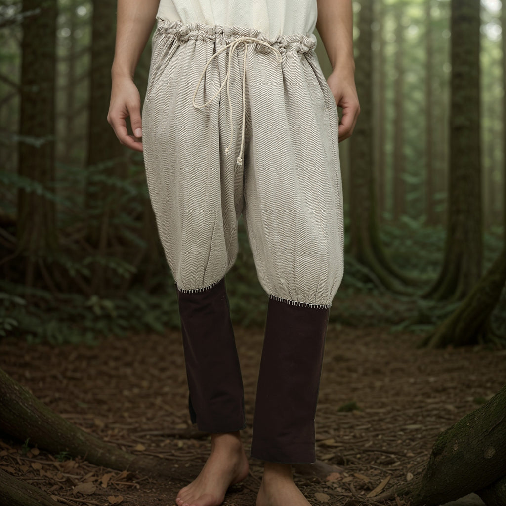 A person stands barefoot in a forest, wearing Nordic Nomad Viking Pants—men’s drawstring trousers with light fabric and contrasting dark knees, inspired by Nordic clothing and ideal for historical reenactment. The upper body is partially out of frame.