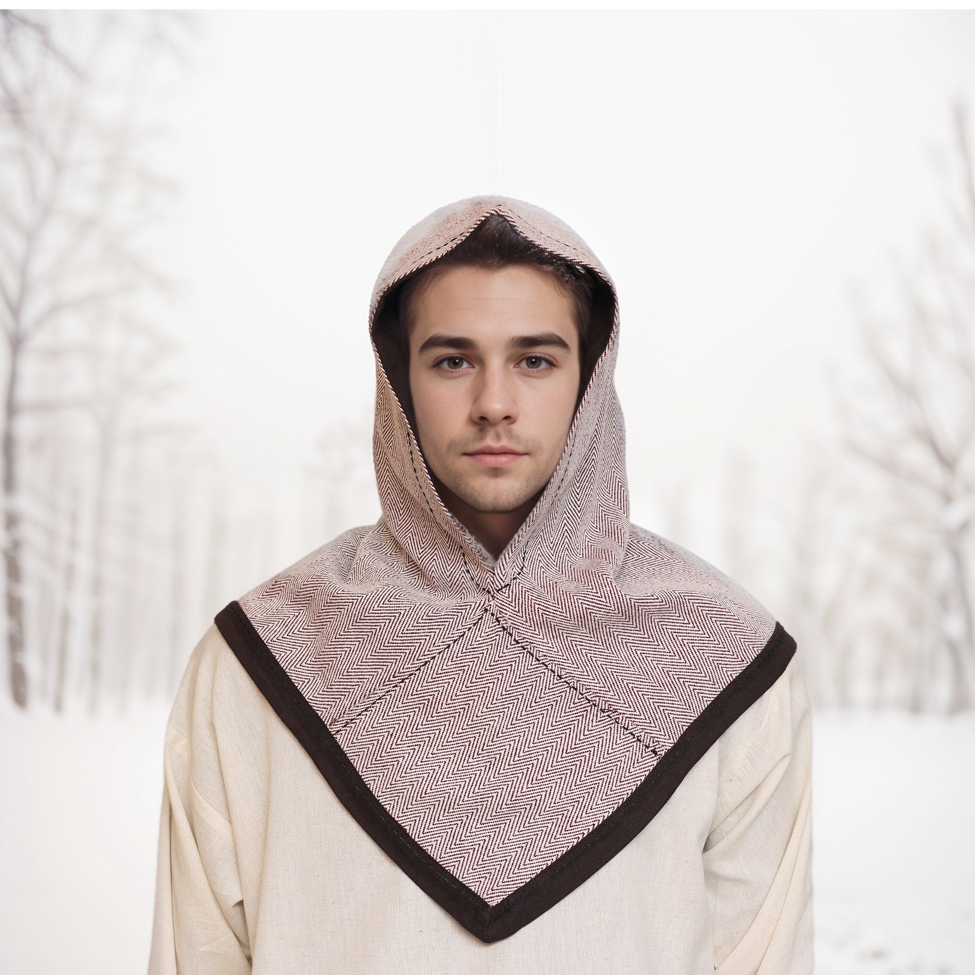 A young man wears the Reversible Viking Hood, Skjoldehamn, with brown trim, standing before a softly blurred snowy forest—an ideal accessory for medieval reenactment.