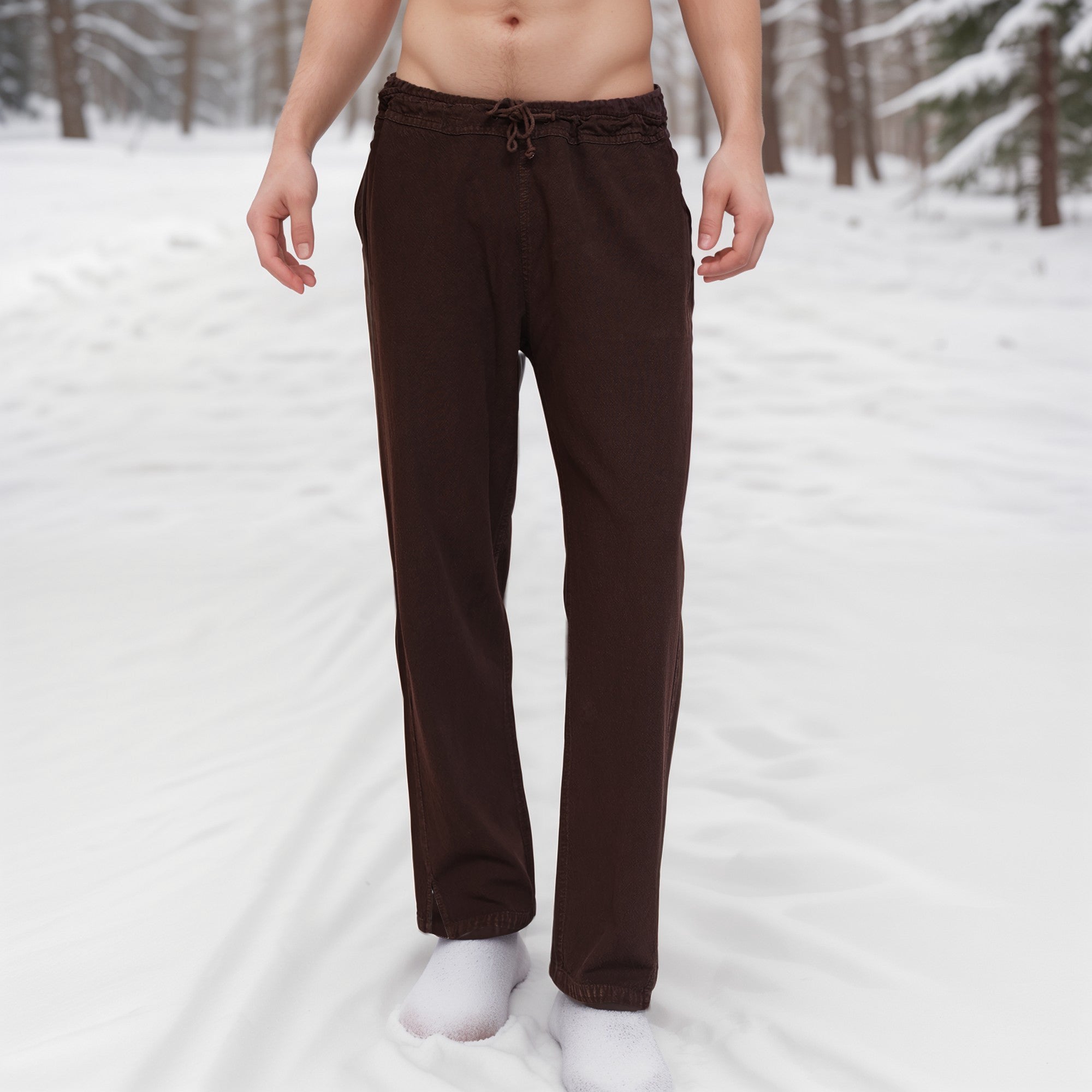 A person stands barefoot in the snow, wearing Warrior Pants- Men's, surrounded by snow-covered trees in a winter forest. Their upper body and face are not visible.