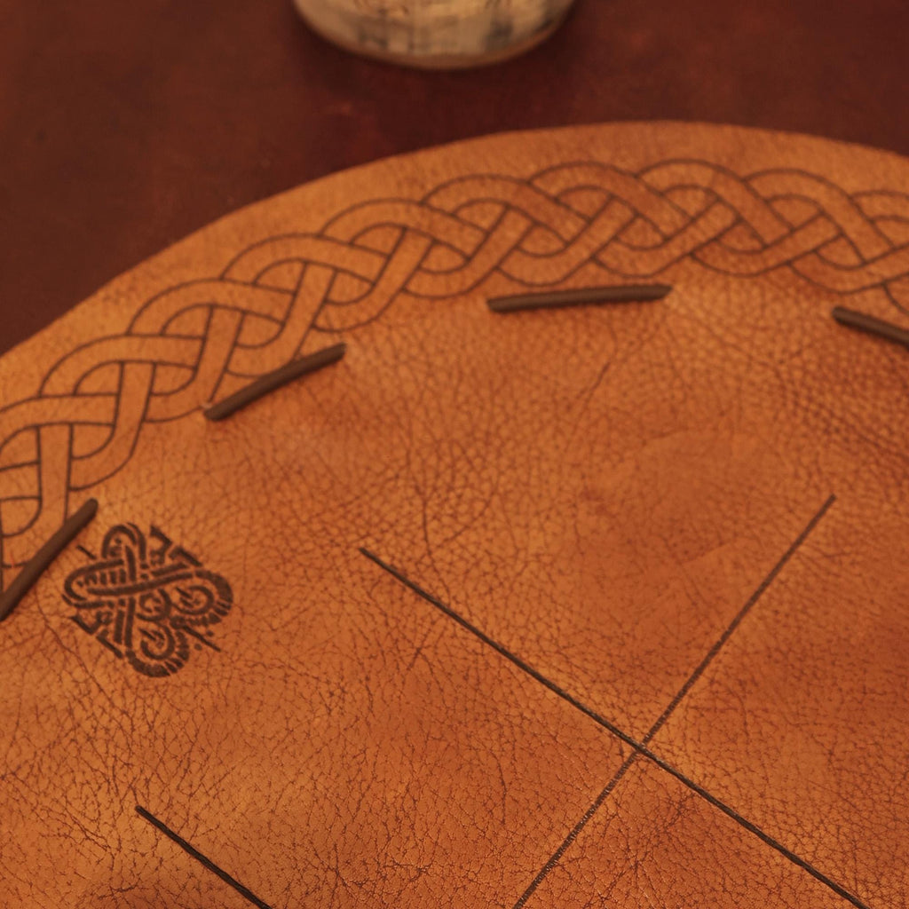 The Viking Leather Tic Tac Toe Game features a circular leather board with a Celtic knot pattern and engraved lines, resembling a handcrafted Viking game. Small wooden sticks are used as markers on the board.
