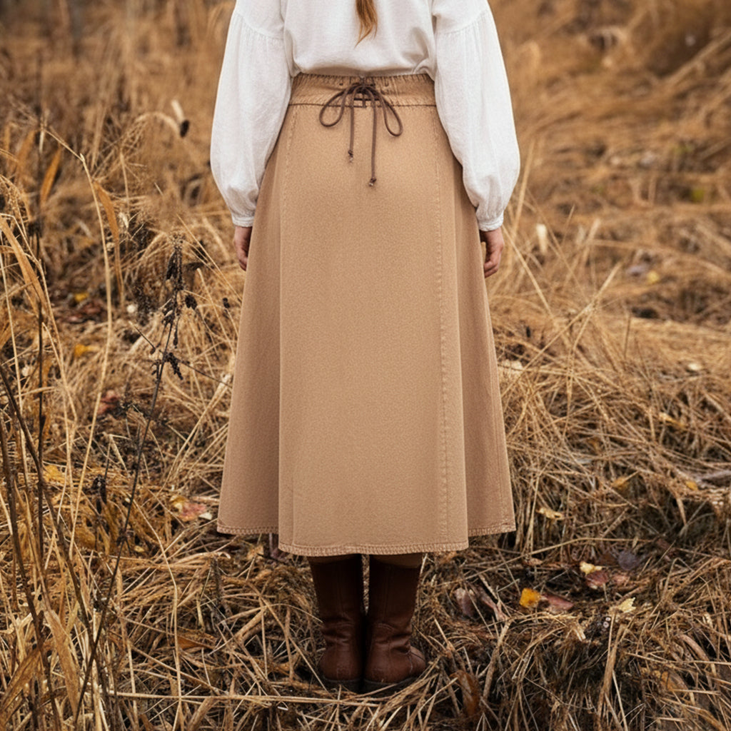 A person with long blonde hair, wearing a white blouse, the Medieval Skirt- Ladies with a tied waist in tan, and brown boots stands in a dry, grassy field facing away from the camera.