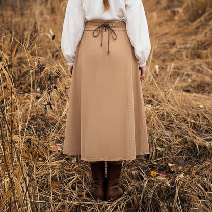 A person with long blonde hair, wearing a white blouse, the Medieval Skirt- Ladies with a tied waist in tan, and brown boots stands in a dry, grassy field facing away from the camera.