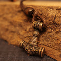 A close-up of a Brass cast Viking Hammer Pendant, adorned with intricate Norse designs, hangs from a braided leather cord and rests against a rugged wooden surface.