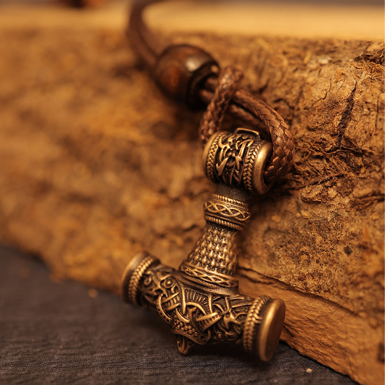 A close-up of a Brass cast Viking Hammer Pendant, adorned with intricate Norse designs, hangs from a braided leather cord and rests against a rugged wooden surface.