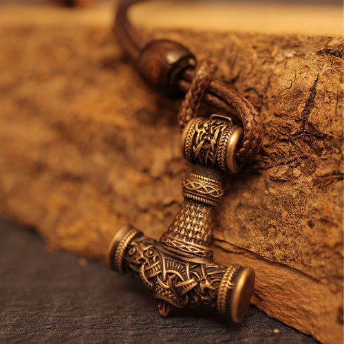 A close-up of a Brass cast Viking Hammer Pendant, adorned with intricate Norse designs, hangs from a braided leather cord and rests against a rugged wooden surface.