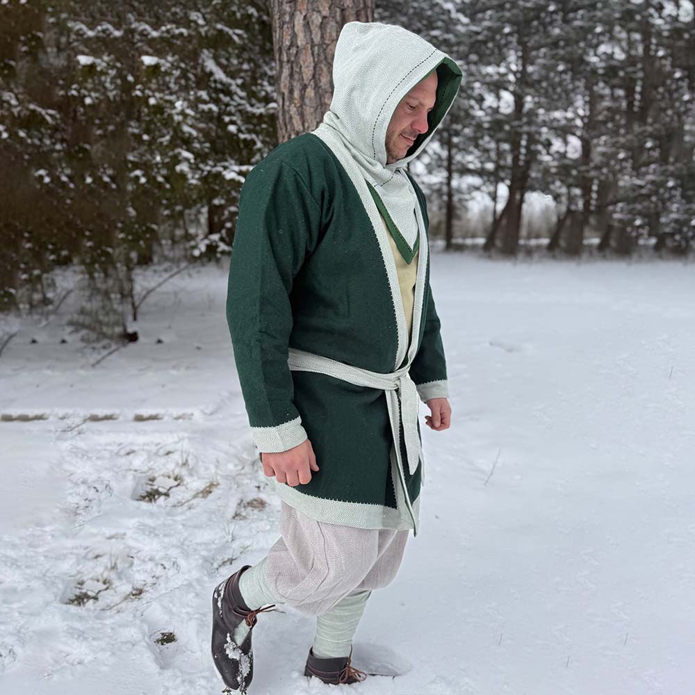 Wearing a green and light gray Viking Wool Coat for men, light pants, and dark boots, a person stands on snowy ground near trees, smiling and looking down.