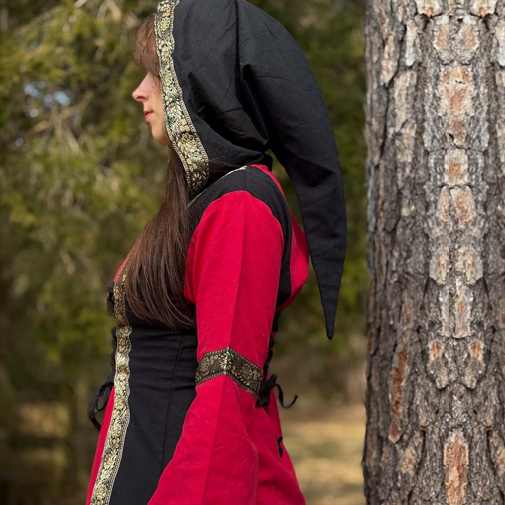 A person in profile stands next to a tree outdoors, wearing the Dark Majesty Medieval Dress—an elegant red and black hooded garment with gold trim, ideal LARP attire for any Renaissance fair. Blurred greenery forms the background.