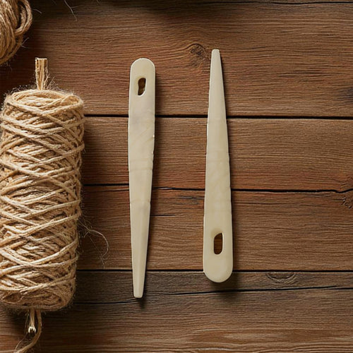 A pair of Bone Knitting Needles – Handcrafted Viking Style – features ivory-colored shafts with oval holes, lying parallel on a wooden surface beside a spool of coarse twine.